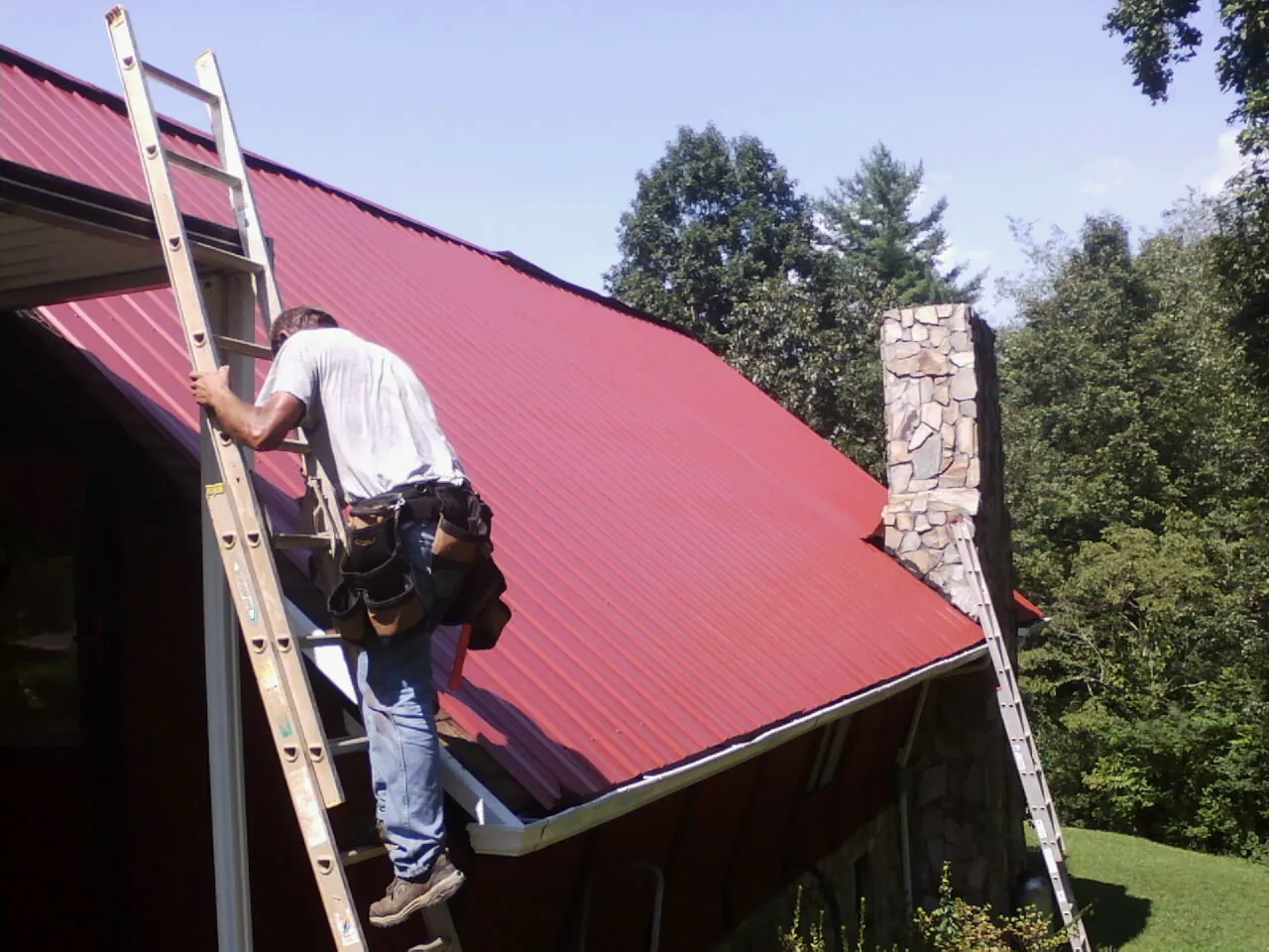 Roofer on a ladder beside a red metal roof and stone chimney — Soffit & Fascia Repair by Roofing Brothers in Helix